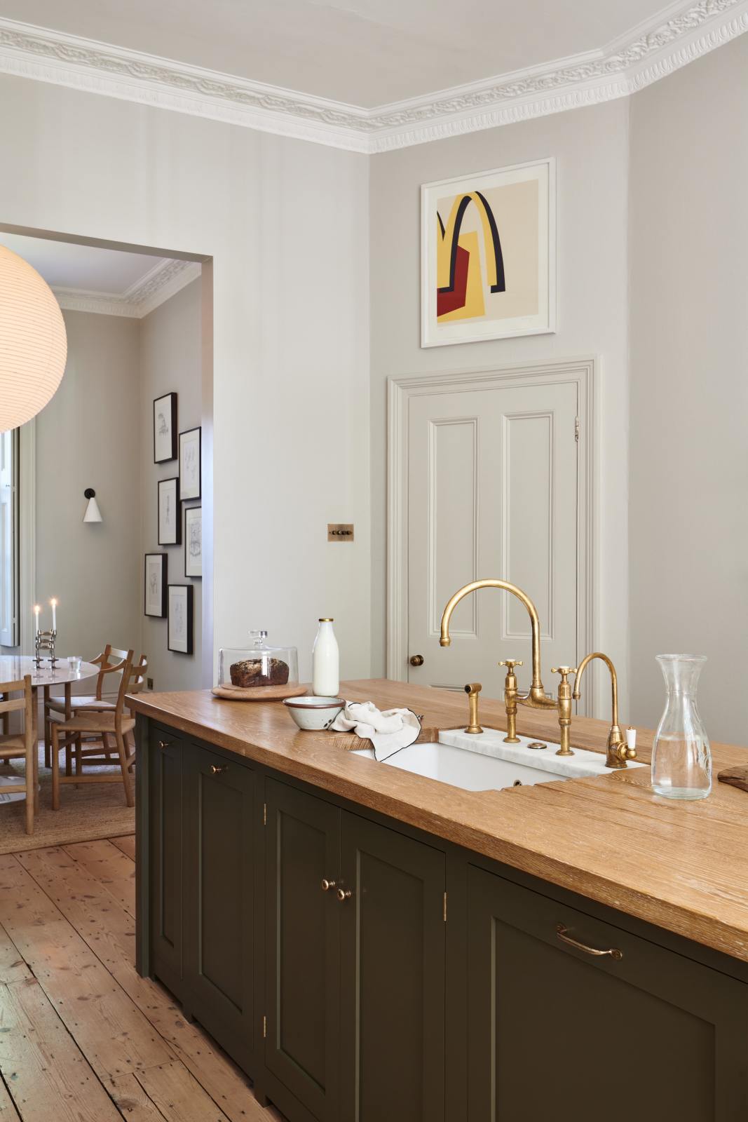 An oblique view of an elegant olive-brown classic kitchen features a kitchen island with a sink, a solid oak worktop, and an aged brass Ionian tap. Beyond the archway lies a separate dining room with a circular wooden table, chairs, and a series of black-framed pencil drawings on the wall.