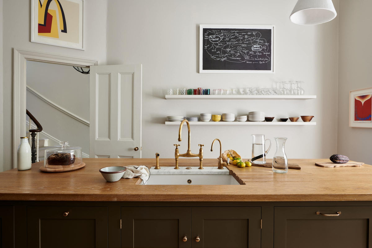 An elegant olive-brown classic kitchen features a kitchen island with a sink, a solid oak worktop, an aged brass Ionian tap, and a breakfast nook with wooden stools at the far edge. Long shelves line the wall, stacked with ceramic bowls, wine glasses, cocktail glasses, and drinking glasses. The tall-ceilinged room is decorated with slender water pitchers, glass milk, a bag of oranges, and big and colourful framed illustrations.