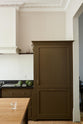 An elegant olive-brown classic kitchen features a cooker run topped with Belgian blue limestone, a kitchen island with a sink and a solid oak worktop, a shaker canopy, and a large full-height fridge/freezer cabinet. The tall-ceilinged room is decorated with slender bottles of wine, vibrant green artichokes, and a glass of water.