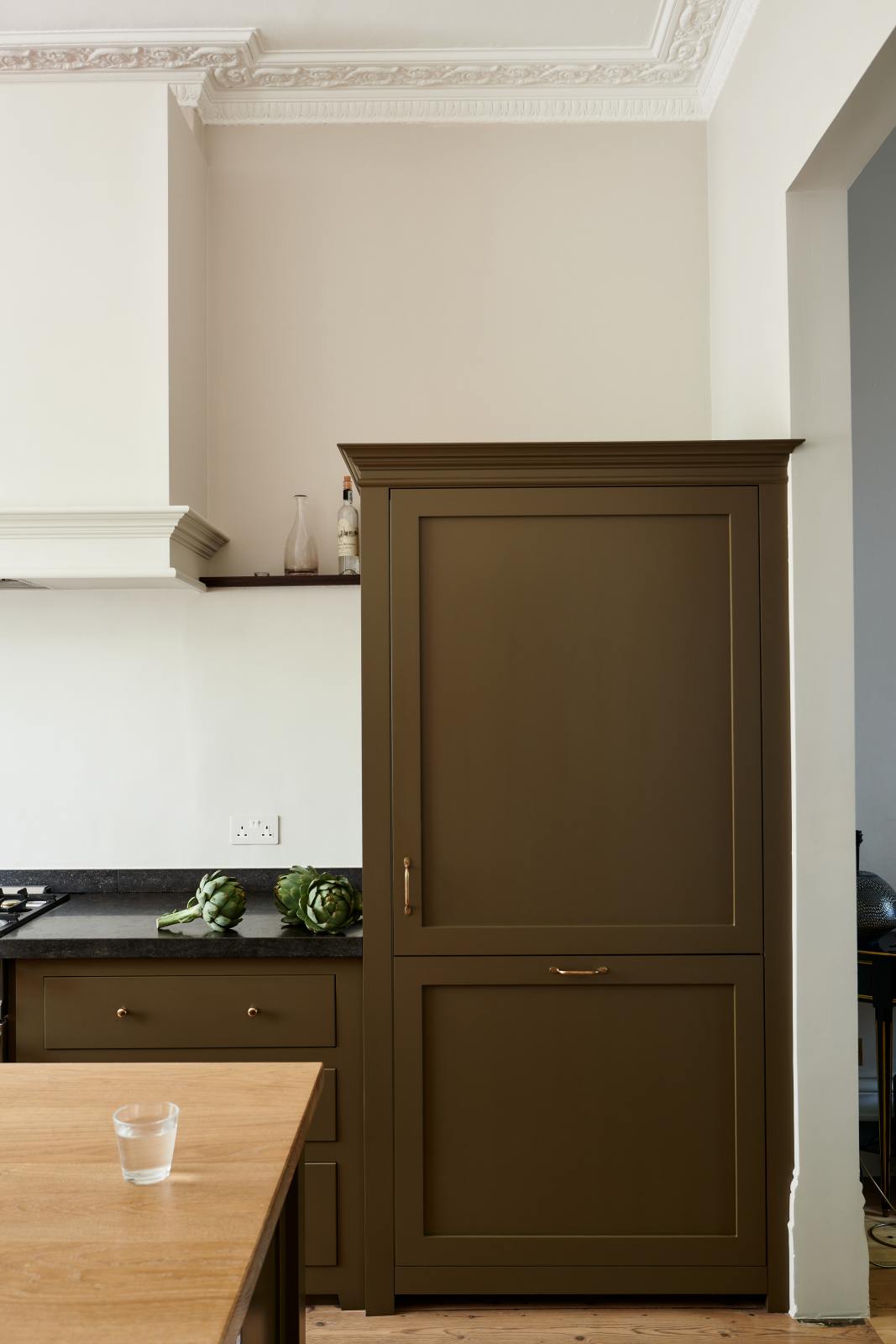 An elegant olive-brown classic kitchen features a cooker run topped with Belgian blue limestone, a kitchen island with a sink and a solid oak worktop, a shaker canopy, and a large full-height fridge/freezer cabinet. The tall-ceilinged room is decorated with slender bottles of wine, vibrant green artichokes, and a glass of water.