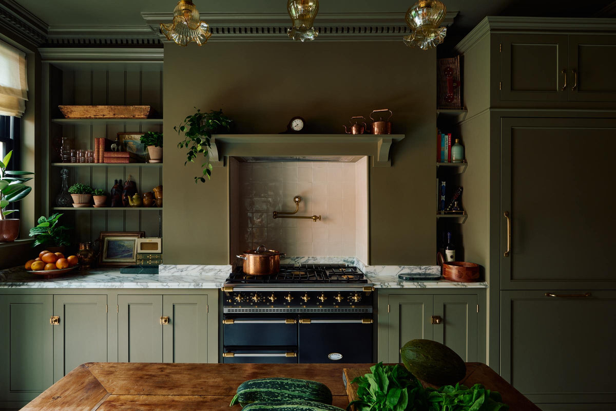 A grand and atmospheric olive green classic English kitchen features a cooker run built around the chimney breast, an antique prep table on castors, a full-height fridge/freezer cabinet with top cupboard and an open countertop cupboard filled with beautiful ceramic mugs, a teapot, potted plants, gold-foiled books, glasses, and framed artwork. The black gas cooker in the overmantle has cream subway splashback tiles and a deVOL pot filler tap.