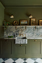 A grand and atmospheric olive green classic English kitchen features a marble worktop, splashback, and shelf along the sink run, a single fluted marble sink and an aged brass Ionian tap. Two beautiful Heirloom wall lights hang above gold-framed famous paintings and potted plants.