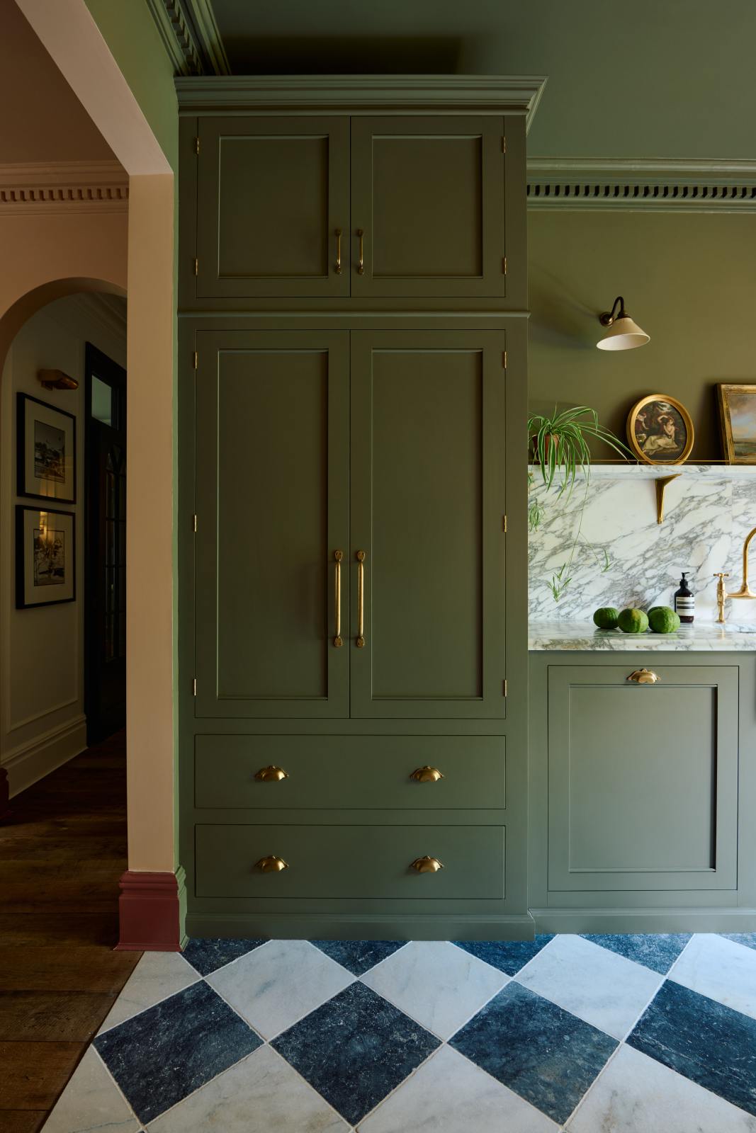 A grand and atmospheric olive green classic English kitchen features a marble worktop, splashback, and shelf along the sink run, a single fluted marble sink and an aged brass Ionian tap. A full-height pantry cabinet takes centre stage, while beside it, beautiful Heirloom wall lights hang above gold-framed paintings and potted plants.