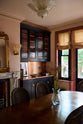 An oblique view of a grand dining room with a printer’s black, classic English café-style nook, reeded glazed cupboards, and aged copper worktops and splashback. A gilded mirror rests above a marble-framed fireplace, framed by amber-frilled sconces.