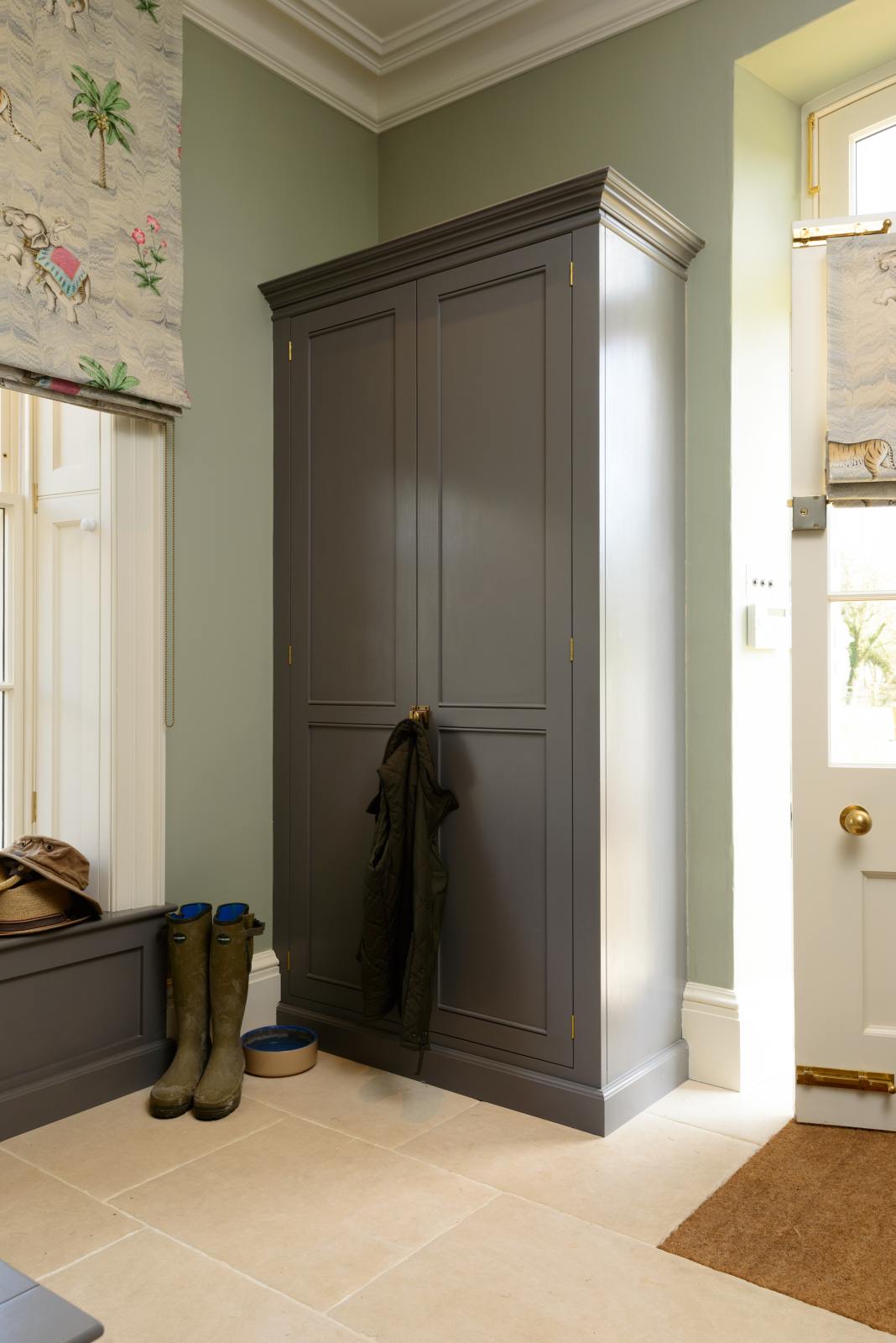 A quiet corner of a functional, muted-grey boot room features an L-shaped bench and a full-height storage cupboard. Beautifully decorated with thick coats, a stack of hats, and a pair of muddy boots, it creates a practical and family-oriented space.