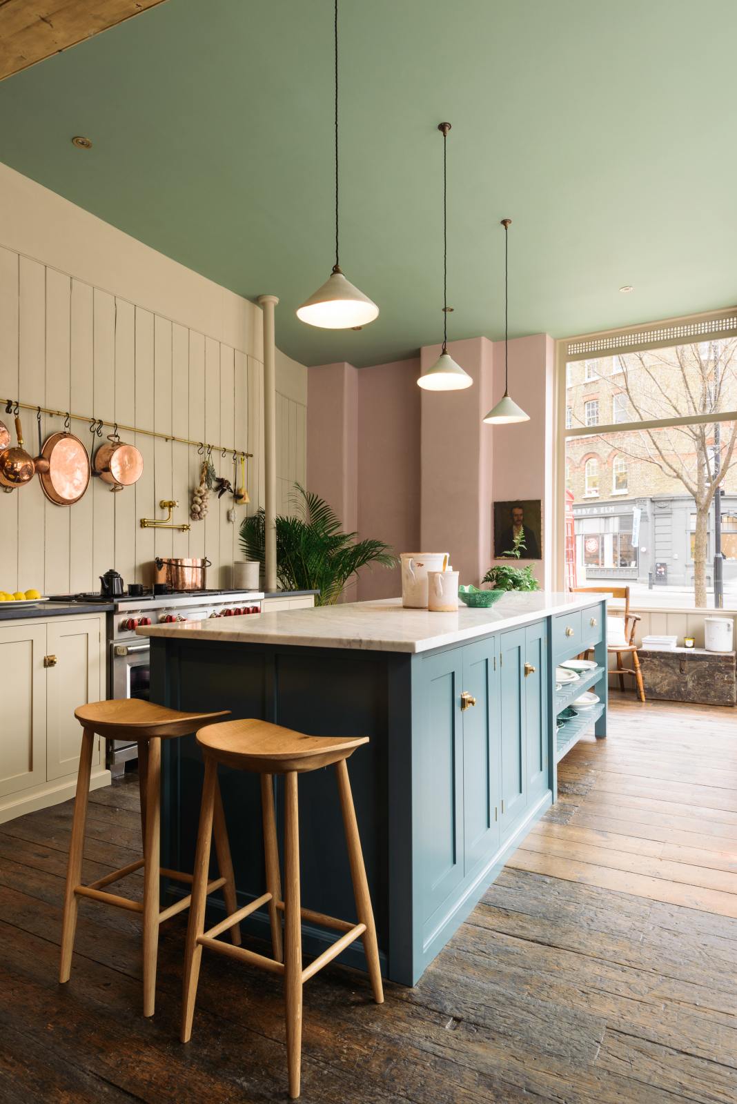 The playful pastel yet classic kitchen features a mushroom-coloured cooker run, aged brass hanging rails, and a pot filler. A long kitchen island with a breakfast nook and deVOL bum stools adds charm. Decorated with copper pots, vintage ceramics, and fresh produce, this kitchen has both functionality and personality.