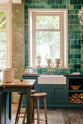 The playful pastel yet classic kitchen features a Clerkenwell blue sink run with a black granite worktop, emerald green splashback tiles, a single fluted Belfast sink, and an aged brass Ionian tap. A long kitchen island with a breakfast nook and deVOL bum stools adds charm. Decorated with chopped firewood, vintage ceramics, and a crystal chandelier, this kitchen has both functionality and personality.