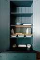 A close-up of a large Clerkenwell blue full-height pantry cupboard with a black granite worktop, fixed shelves, a wooden dish rack, and a variety of ceramic plates, dishes and colanders.