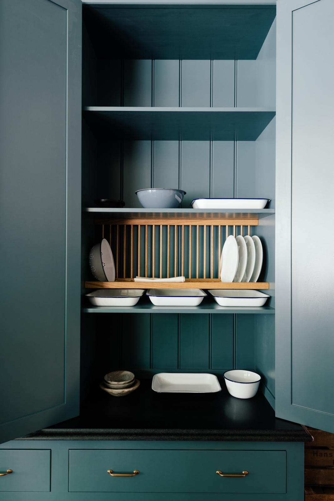 A close-up of a large Clerkenwell blue full-height pantry cupboard with a black granite worktop, fixed shelves, a wooden dish rack, and a variety of ceramic plates, dishes and colanders.
