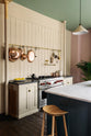 An oblique view of a beige cooker run with a black granite worktop, a silver gas cooker, an aged brass hanging rail, an aged brass pot filler and a long kitchen island with a Carrara marble worktop. Lovingly decorated with hanging copper pots and pans, fresh lemons, garlic, chilli and sage, this space balances functionality and style.