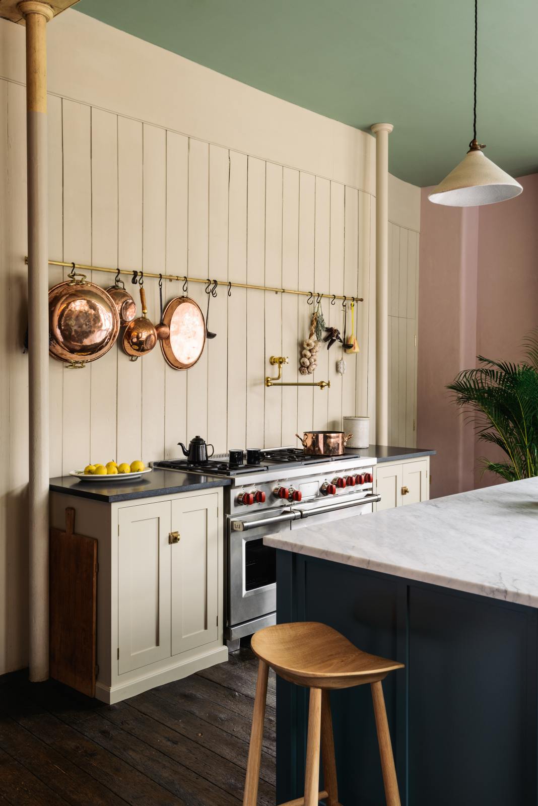 An oblique view of a beige cooker run with a black granite worktop, a silver gas cooker, an aged brass hanging rail, an aged brass pot filler and a long kitchen island with a Carrara marble worktop. Lovingly decorated with hanging copper pots and pans, fresh lemons, garlic, chilli and sage, this space balances functionality and style.