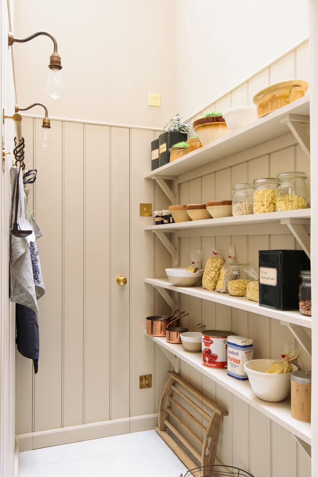 A narrow, high-ceilinged pantry painted beige, featuring four shelves filled with dried ingredients, copper pots and pans, jars of jam, a large can of tomato paste, and an aged brass hanging rail.