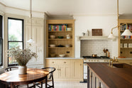 A lovely and simple blush yellow, classic English kitchen features a cooker run with Carrara marble worktop, two open countertop cupboards, a white gas stove inside a chimney breast, porcelain splashback, a purple-brown kitchen island with a solid oak worktop and aged brass Mayan taps. A circular coffee table with matching chairs, a deVOL gaselier light, and a collection of muted ceramics and tall wildflowers create an elegant and inviting atmosphere in the room.