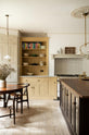 A lovely and simple blush yellow, classic English kitchen features a cooker run with Carrara marble worktop, two open countertop cupboards, a white gas stove inside a chimney breast, porcelain splashback, a purple-brown kitchen island with a solid oak worktop and aged brass Mayan taps. A circular coffee table with matching chairs, a deVOL gaselier light, and a collection of muted ceramics and tall wildflowers create an elegant and inviting atmosphere in the room.