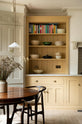A lovely and simple blush yellow, classic English kitchen features a cooker run with Carrara marble worktop, two open countertop cupboards, a white gas stove inside a chimney breast, porcelain splashback, a purple-brown kitchen island with a solid oak worktop and aged brass Mayan taps. A circular coffee table with matching chairs, and a collection of muted ceramics and tall wildflowers create an elegant and inviting atmosphere in the room.