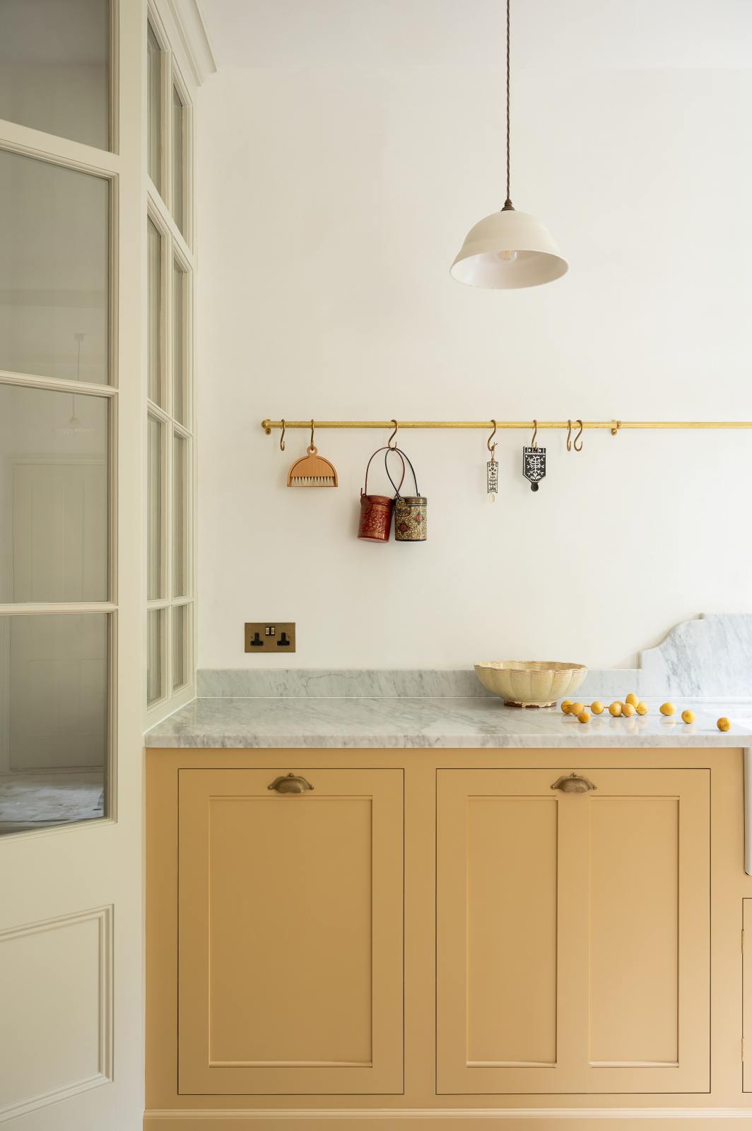 A charming blush yellow, classic English kitchen features a Carrara marble countertop, an ornate backsplash, and a porcelain pendant light. Decorated with copper pots, vintage ceramics, and small wooden cleaning supplies, it creates an elegant and tidy atmosphere.