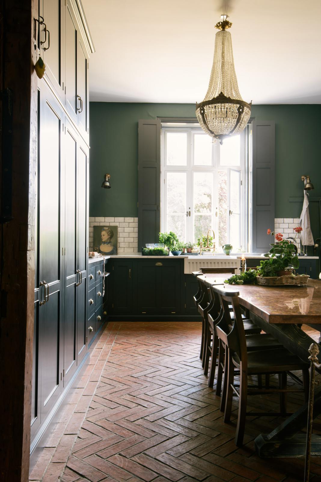 A grand L-shaped kitchen with bespoke Georgian-style cabinetry, painted in a pantry blue and decorated with traditional brass fittings. The room has a high ceiling and tall painted shutters on either side of a beautiful original window. The walls are a soft moss green with a band of mottled white metro tiles. A large antique crystal chandelier hangs above a rustic herringbone brick floor.