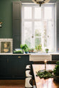A grand L-shaped kitchen with bespoke Georgian-style cabinetry, painted in a pantry blue and decorated with traditional brass fittings. The room has a high ceiling and tall painted shutters on either side of a beautiful original window. The walls are a soft moss green with a band of mottled white metro tiles. A large antique crystal chandelier hangs above a rustic herringbone brick floor.