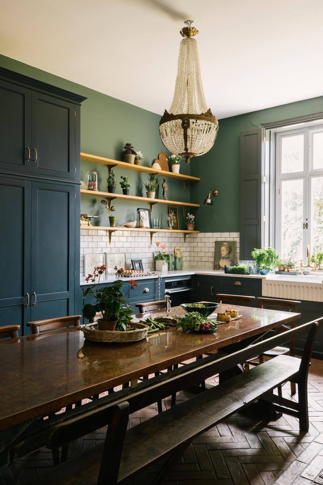 The spacious kitchen in the renovated rectory features a blue L-shaped countertop, a mix of cabinetry against green walls, and a metro tile backsplash. At the centre, a copper-topped table flanked by a vintage bench and chairs is complemented by an elegant antique chandelier.