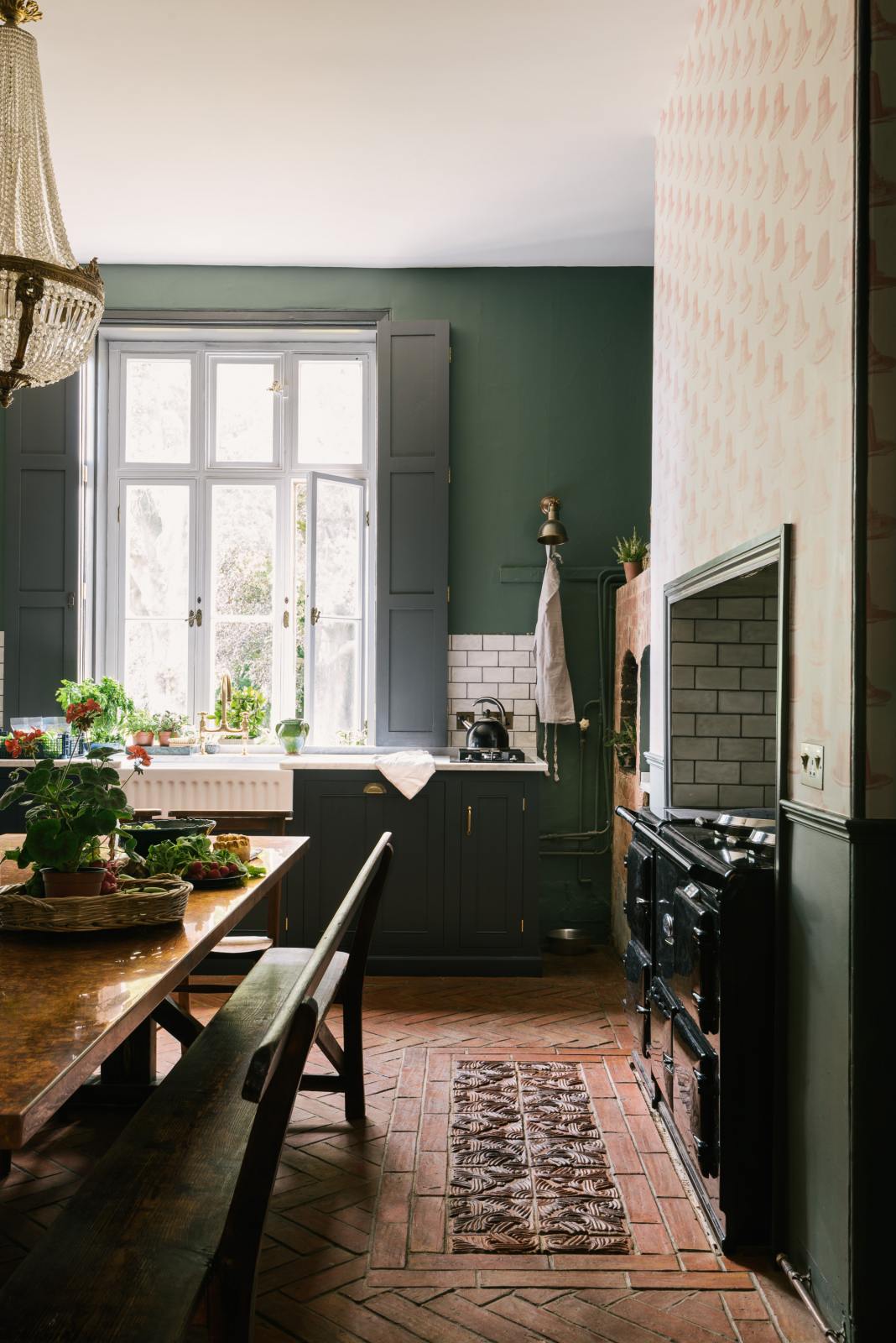 The spacious kitchen in the renovated rectory features a blue L-shaped countertop, a mix of cabinetry against green walls, and a metro tile backsplash. At the centre, a copper-topped table flanked by a vintage bench and chairs is complemented by an elegant antique chandelier.
