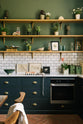 The deep blue base cupboards contain an under-counter oven and are topped with Carrara marble. A lovely white metro tile splashback extends up the wall to meet the first three wooden shelves, each supported by beautiful handmade aged brass brackets. The walls are painted a muted dark green, and the shelves display an array of trinkets, plants, and artwork.