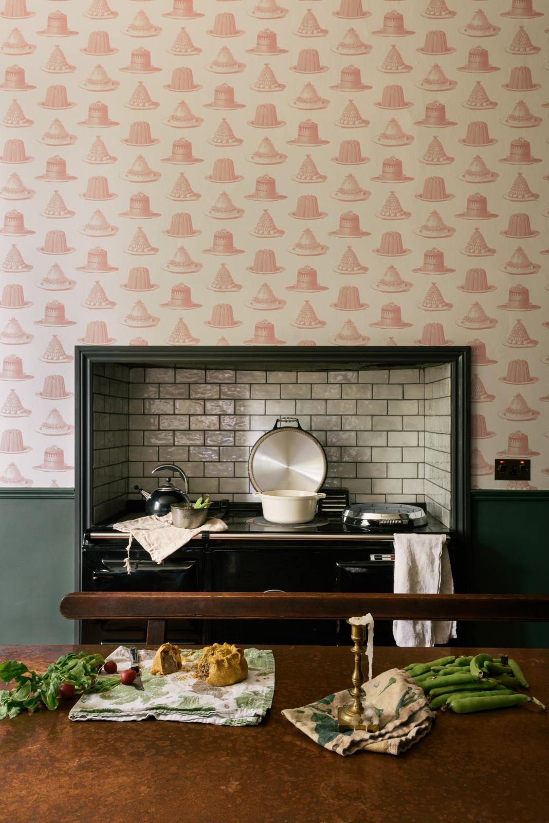 The image features a close-up view of an aged copper tabletop in the foreground. In the background, there is a chimney breast with a black Aga stove situated in the alcove. The chimney breast is adorned with whimsical wallpaper that has a pale cream base colour, decorated with illustrations of light pink jellies and cakes.