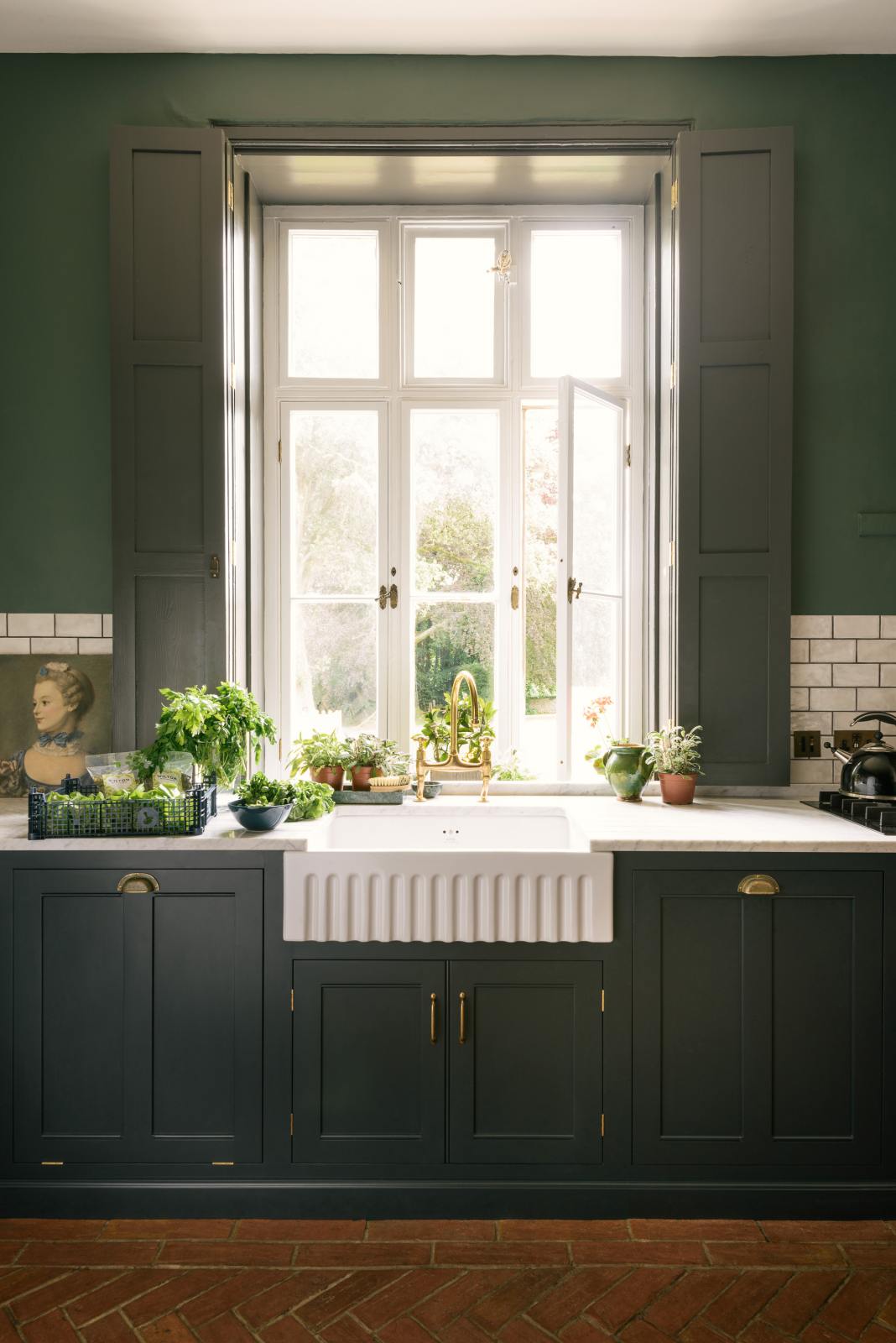 A grand L-shaped kitchen with bespoke Georgian-style cabinetry, painted in a pantry blue and decorated with traditional brass fittings. The room has a high ceiling and tall painted shutters on either side of a beautiful original window. The walls are a soft moss green with a band of mottled white metro tiles. A large antique crystal chandelier hangs above a rustic herringbone brick floor.