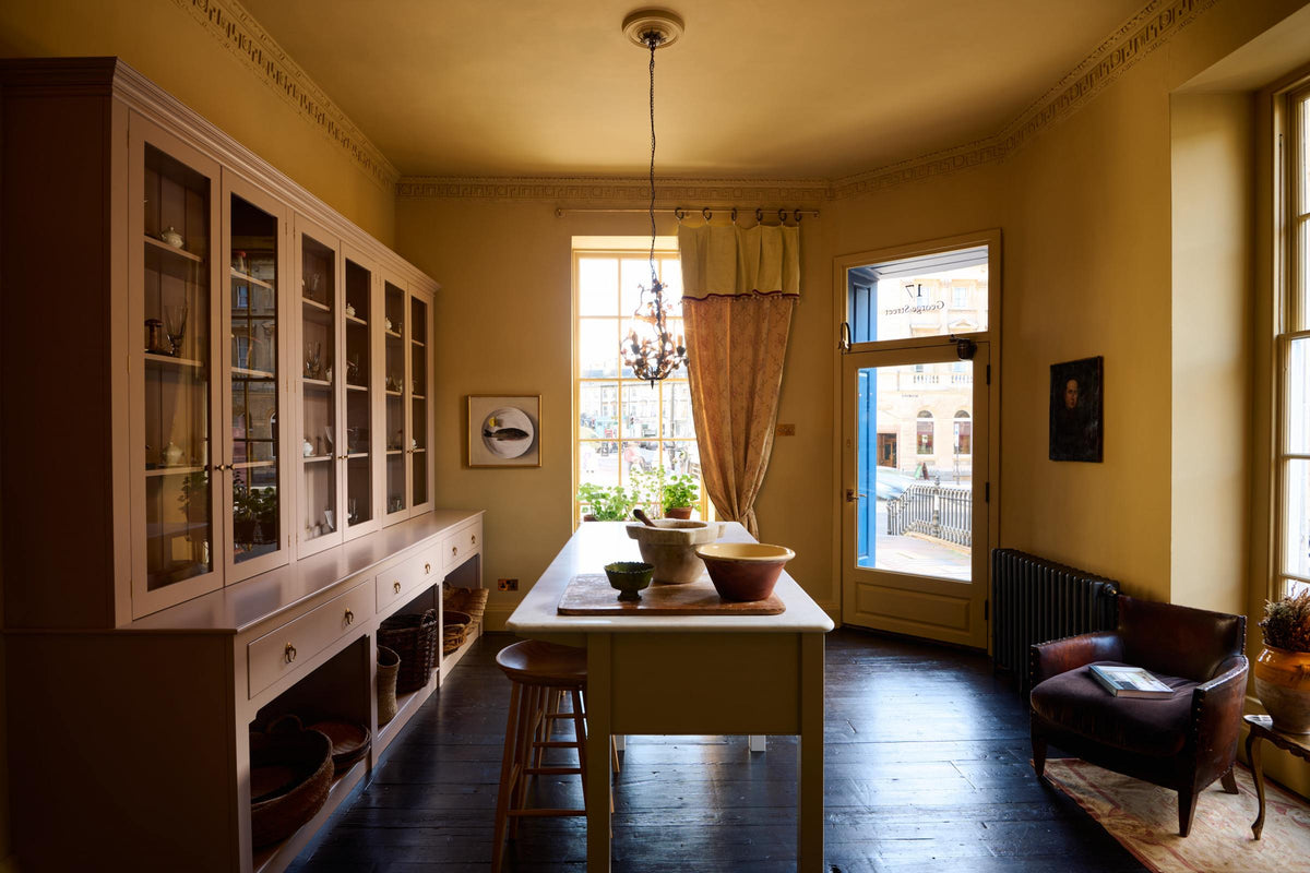 A warm yellow room with a large Princelet pink dresser showcasing glazed cupboards filled with delicate ceramics, papier-mâché mushrooms, and wooden baskets along the bottom, giving the space a relaxed, living-room feel. The space includes a wide prep table with an open nook for deVOL bum stools. There’s also a vintage armchair sofa, large ceramic bowls, a metal vines-and-flowers chandelier, and a golden-framed painting of a fish and a lemon.