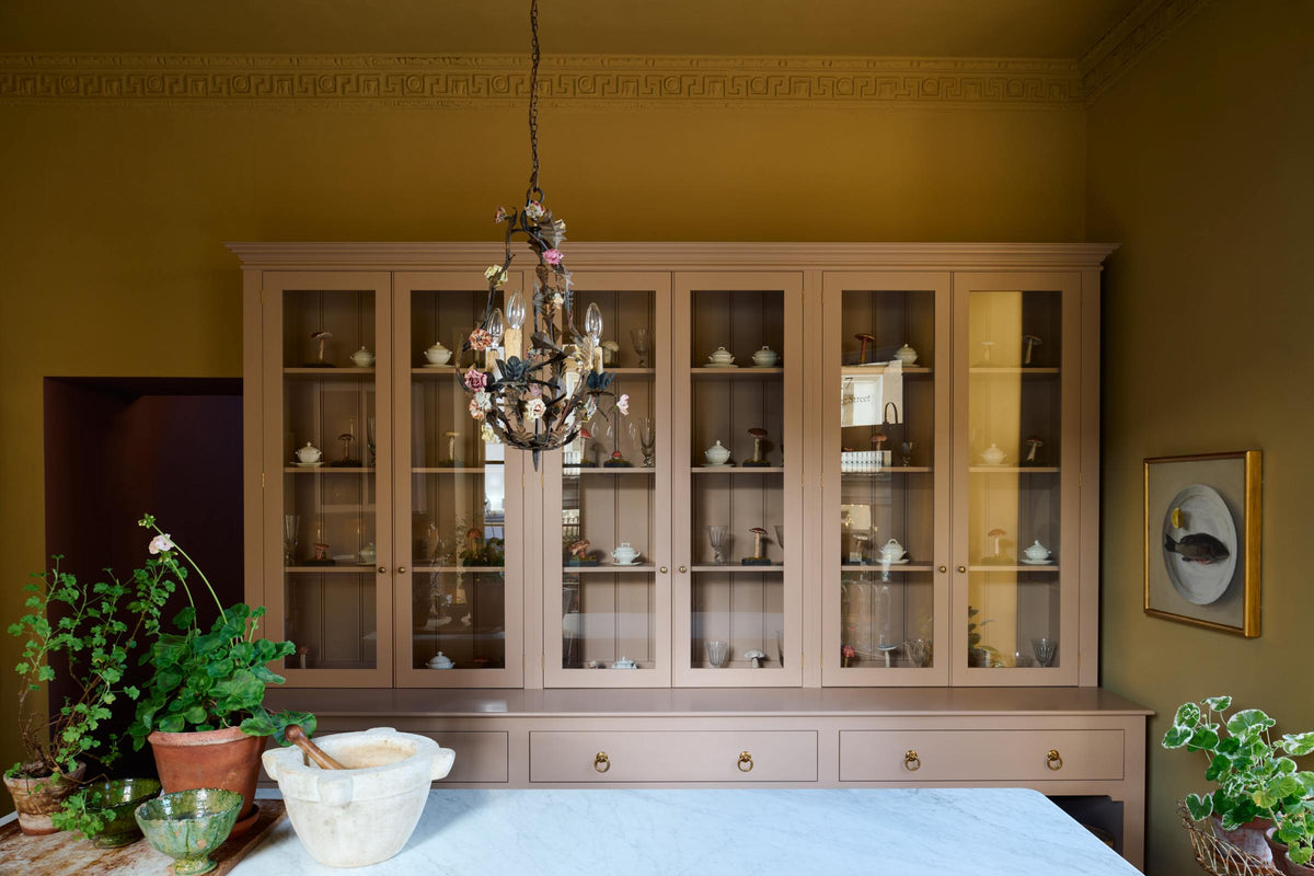 A warm yellow room with a large Princelet pink dresser showcasing glazed cupboards filled with delicate ceramics, drinking glasses, and papier-mâché mushrooms, giving the space a relaxed, living-room feel. The space includes a wide prep table, large ceramic bowls, a metal vines-and-flowers chandelier, and a golden-framed painting of a fish and a lemon.