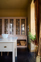 A warm yellow room with a large Princelet pink dresser showcasing glazed cupboards filled with delicate ceramics, drinking glasses, and papier-mâché mushrooms, giving the space a relaxed, living-room feel. The space includes a wide prep table, large ceramic bowls, a metal vines-and-flowers chandelier, and a golden-framed painting of a fish and a lemon.