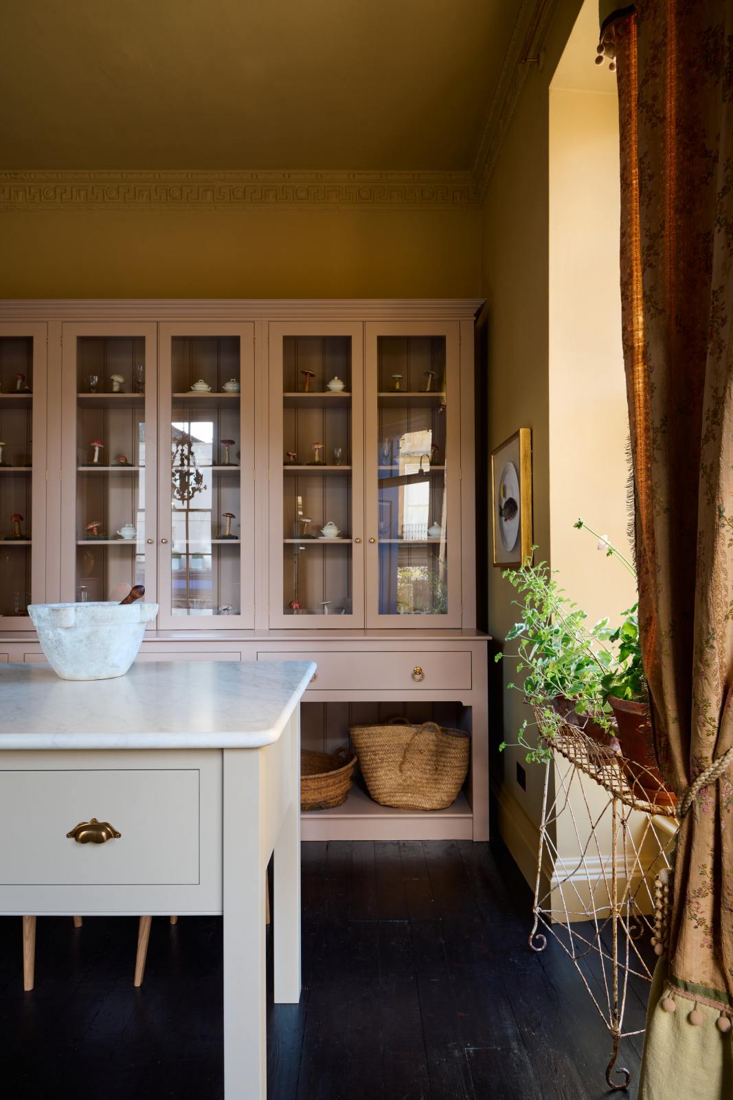 A warm yellow room with a large Princelet pink dresser showcasing glazed cupboards filled with delicate ceramics, drinking glasses, and papier-mâché mushrooms, giving the space a relaxed, living-room feel. The space includes a wide prep table, large ceramic bowls, a metal vines-and-flowers chandelier, and a golden-framed painting of a fish and a lemon.