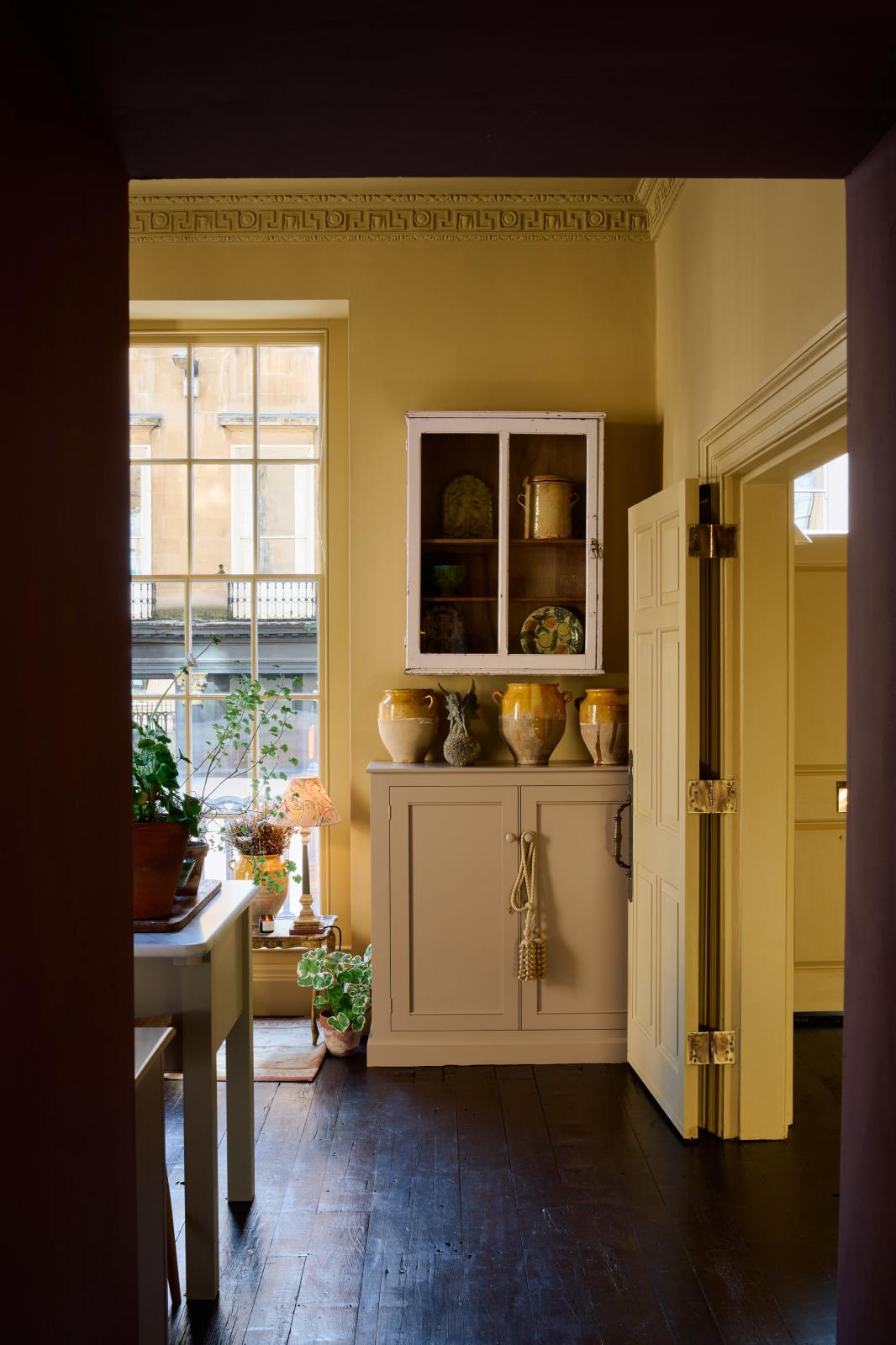 The room is painted a warm yellow and features a wide prep table with an open nook for deVOL bum stools. A Princelet pink base cupboard adds a cheerful touch, while a vintage white metal wall cupboard provides additional storage. The space is beautifully decorated with mustard yellow accents, vintage ceramic vases and furniture, and a plate illustrated with citrus fruits. A vintage gold-finish metal side table completes the bohemian style of the room.