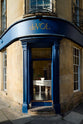 An open front door at the edge of a traditional Bath building, outlined with blue frames and featuring a golden deVOL logo at the centre. Inside is a sneak preview of a warm yellow room with a wide, linen white prep table, a large Princelet pink dresser showcasing glazed cupboards filled with delicate ceramics,  and a metal vines-and-flowers chandelier, giving the space a relaxed, living-room feel.