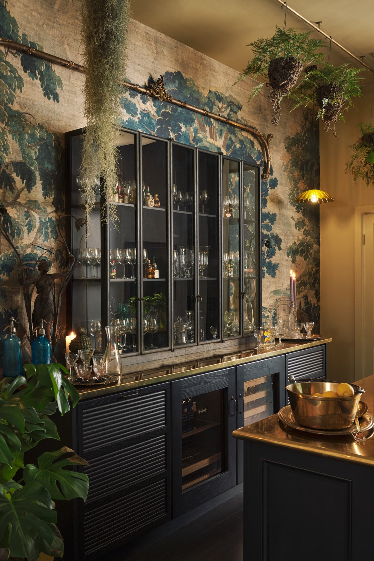 An oblique view of a chic and glamorous Bobbin Black Haberdasher’s bar featuring a kitchen island with a polished brass worktop, glazed base cabinet with internal drawers, and tall countertop cupboards. Set against an illustrated foliage wallpaper and adorned with hanging plants, wine glasses, and vintage decanters, it creates a beautiful and intimate personal bar.