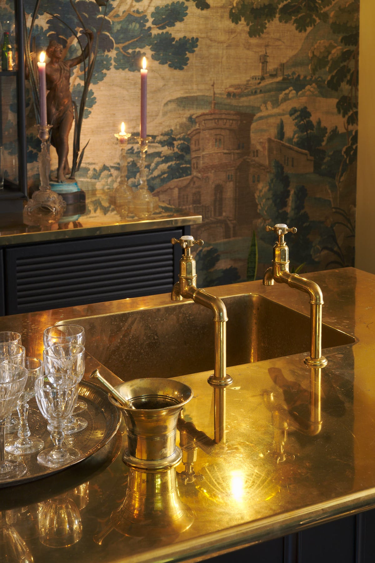 A close-up of a chic and glamorous Bobbin Black Haberdasher’s bar featuring a kitchen island with a polished brass worktop, a single polished brass sink, aged brass Mayan taps, glazed base cabinets with internal drawers, and tall countertop cupboards. Set against an illustrated foliage wallpaper and adorned with wine glasses, an antique brass figurine, crystal handle holders, trays, and vintage decanters, it creates a beautiful and intimate personal bar.