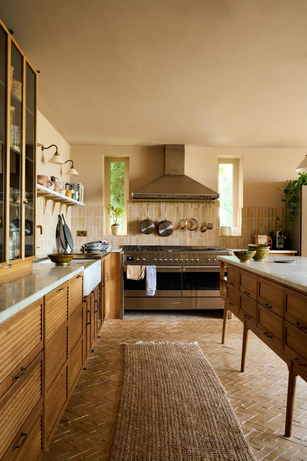Nestled between a warm and chic Haberdasher’s kitchen features an L-shaped sink and cooker run, countertop cupboards, a kitchen island, a Carrara and copper mix worktop, and a double-fluted marble sink, is a large stainless steel gas cooker with a matching canopy. Adorned with artisan ceramics, Heirloom wall lights, a large vintage rug, and luscious green house plants, this kitchen creates a warm and inviting Spanish-inspired space in Yorkshire.