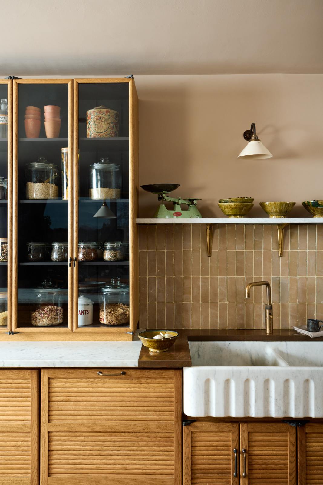 A quiet corner of a warm and chic Haberdasher’s kitchen featuring an L-shaped sink and cooker run, countertop cupboards, a Carrara and copper mix worktop, and a double-fluted marble sink. Adorned with artisan ceramics, Heirloom wall lights, dried ingredients in glass jars, and a vintage green scale, this kitchen creates a warm and inviting Spanish-inspired space in Yorkshire.