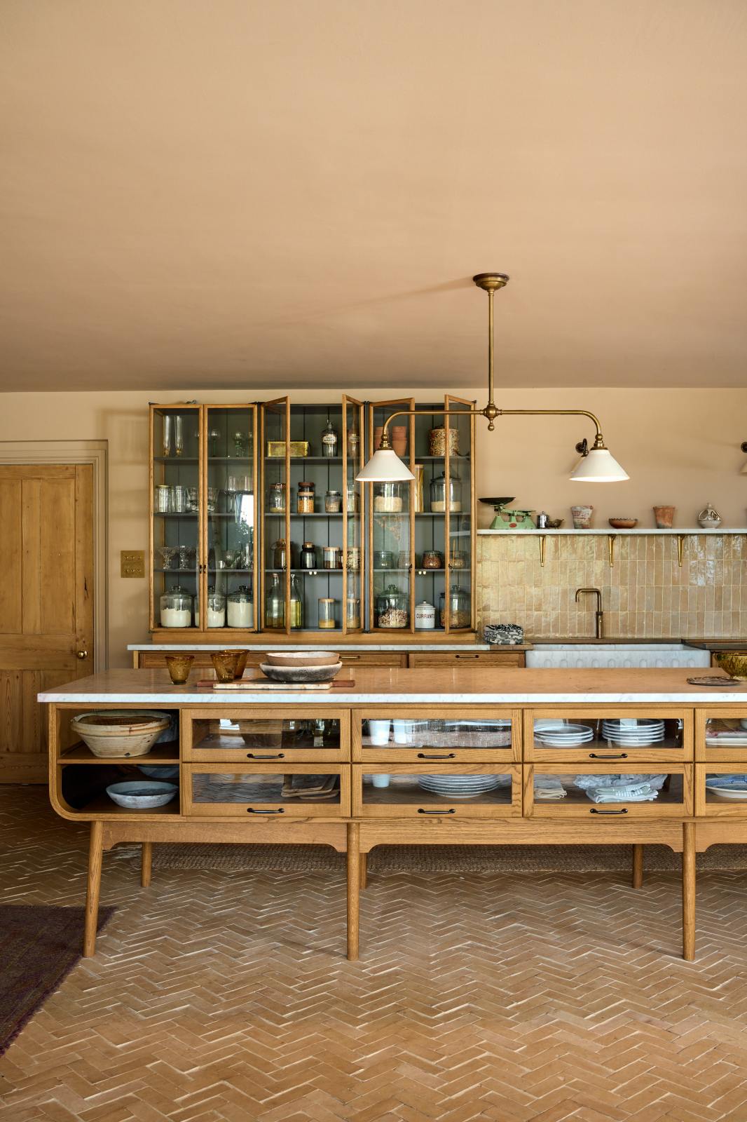 A warm and chic Haberdasher’s kitchen features an L-shaped sink and cooker run, countertop cupboards, a kitchen island, a Carrara and copper mix worktop, and a double-fluted marble sink. Adorned with artisan ceramics, a deVOL gaselier light, Heirloom wall lights, large vintage rugs, and engraved drinking glasses, this kitchen creates a warm and inviting Spanish-inspired space in Yorkshire.