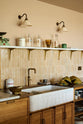 A close-up of a warm and chic Haberdasher’s kitchen features an L-shaped sink and cooker run, countertop cupboards, a Carrara and copper mix worktop, and a double-fluted marble sink. Adorned with artisan ceramics, Heirloom wall lights, dried ingredients in glass jars, and a vintage green scale, this kitchen creates a warm and inviting Spanish-inspired space in Yorkshire.