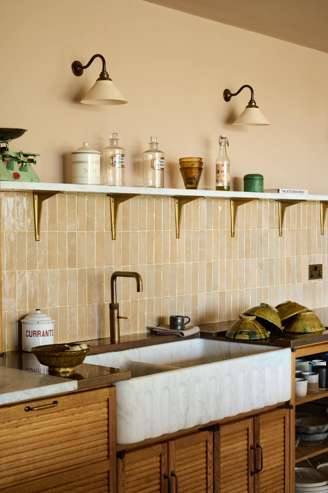 A close-up of a warm and chic Haberdasher’s kitchen features an L-shaped sink and cooker run, countertop cupboards, a Carrara and copper mix worktop, and a double-fluted marble sink. Adorned with artisan ceramics, Heirloom wall lights, dried ingredients in glass jars, and a vintage green scale, this kitchen creates a warm and inviting Spanish-inspired space in Yorkshire.