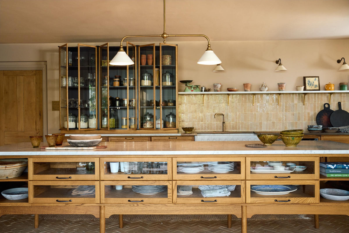 A warm and chic Haberdasher’s kitchen features an L-shaped sink and cooker run, countertop cupboards, a kitchen island, a Carrara and copper mix worktop, and a double-fluted marble sink. Adorned with artisan ceramics, a deVOL gaselier light, Heirloom wall lights, large vintage rugs, and engraved drinking glasses, this kitchen creates a warm and inviting Spanish-inspired space in Yorkshire.