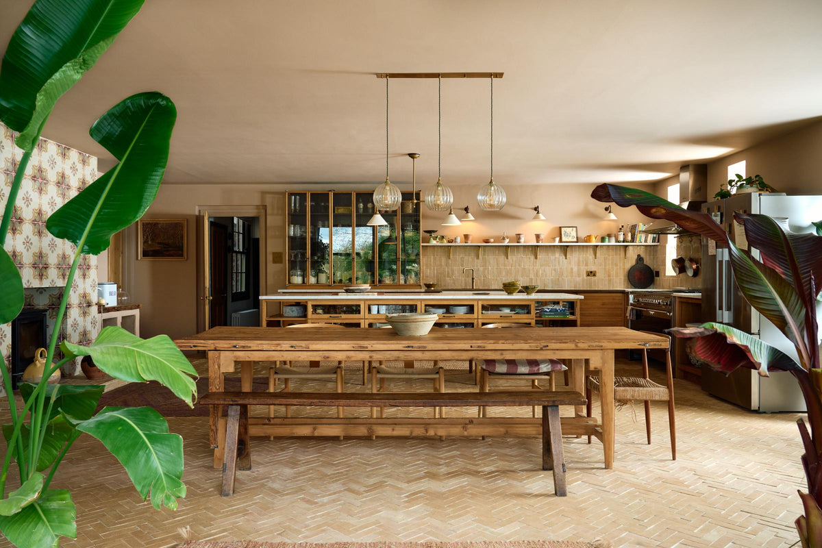 A warm and chic Haberdasher’s kitchen features an L-shaped sink and cooker run, countertop cupboards, a kitchen island, a Carrara and copper mix worktop, and a double-fluted marble sink. Adorned with artisan ceramics, a deVOL gaselier light, Heirloom wall lights, large vintage rugs, and luscious green house plants, this kitchen creates a warm and inviting Spanish-inspired space in Yorkshire.