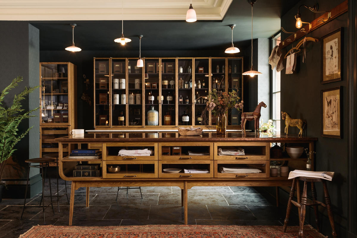 A cool and elegant Haberdasher’s collection features a large prep table with a copper worktop, a pantry run with tambour base cabinets, glazed countertop cabinets, and a freestanding pantry with glazed doors. Adorned with vintage furniture and items, artisan ceramics, the deVOL porcelain light collection, Creedy stools, assorted containers, bottles, and a stunning bouquet of dried flowers, this space creates an atmospheric social bar.