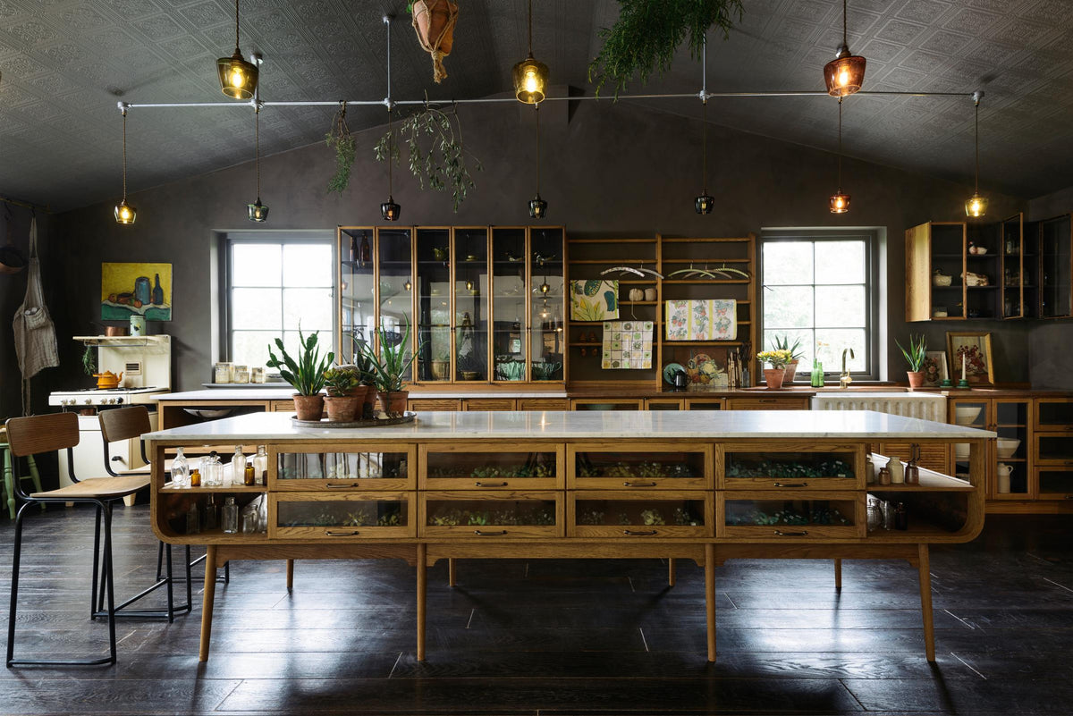 A warm and inviting Haberdasher’s kitchen features a long sink run with a Carrara marble and copper worktop, a double-fluted marble sink, a kitchen island, wall cupboards, and countertop cupboards. Adorned with luscious green hanging and potted plants, vintage items, artisan ceramics, illustrative tea towels, paintings, and a beloved white gas stove, this kitchen creates a lovely exhibitionist space that is both beautiful and practical.