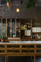 A warm and inviting Haberdasher’s kitchen features a long sink run with a Carrara marble and copper worktop, a double-fluted marble sink, a kitchen island, wall cupboards, and countertop cupboards. Adorned with luscious green hanging and potted plants, vintage items, artisan ceramics, illustrative tea towels, paintings, and a beloved white gas stove, this kitchen creates a lovely exhibitionist space that is both beautiful and practical.