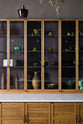 A closer look at a warm and inviting Haberdasher’s kitchen features a long sink run with a Carrara marble and copper worktop, along with countertop cupboards. Adorned with luscious green hanging and potted plants, vintage items, and artisan ceramics, this kitchen creates a lovely exhibitionist space that is both beautiful and practical.