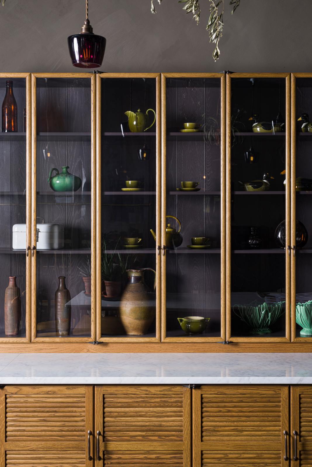A closer look at a warm and inviting Haberdasher’s kitchen features a long sink run with a Carrara marble and copper worktop, along with countertop cupboards. Adorned with luscious green hanging and potted plants, vintage items, and artisan ceramics, this kitchen creates a lovely exhibitionist space that is both beautiful and practical.