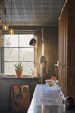 Nestled between a long sink run with a Carrara marble and copper worktop and countertop cupboards, the warm and inviting Haberdasher’s kitchen is adorned with luscious green hanging and potted plants, vintage items, artisan ceramics, illustrative tea towels, paintings, and a beloved white gas stove. This kitchen creates a lovely exhibitionist space that is both beautiful and practical.