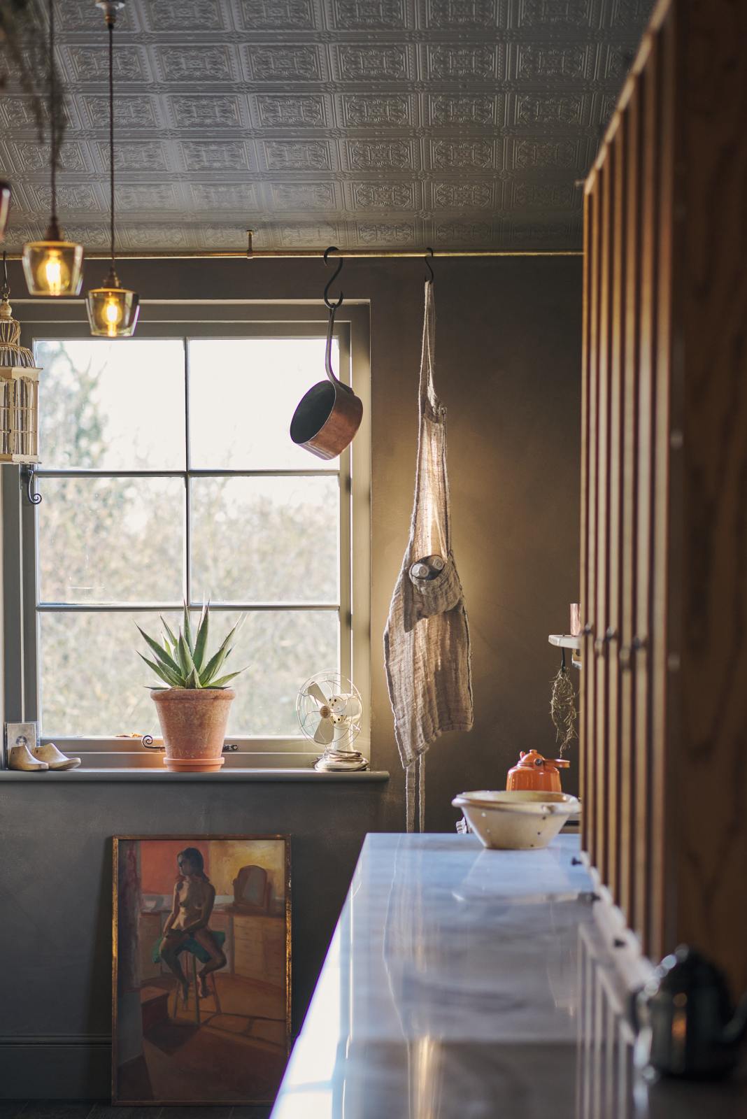 Nestled between a long sink run with a Carrara marble and copper worktop and countertop cupboards, the warm and inviting Haberdasher’s kitchen is adorned with luscious green hanging and potted plants, vintage items, artisan ceramics, illustrative tea towels, paintings, and a beloved white gas stove. This kitchen creates a lovely exhibitionist space that is both beautiful and practical.