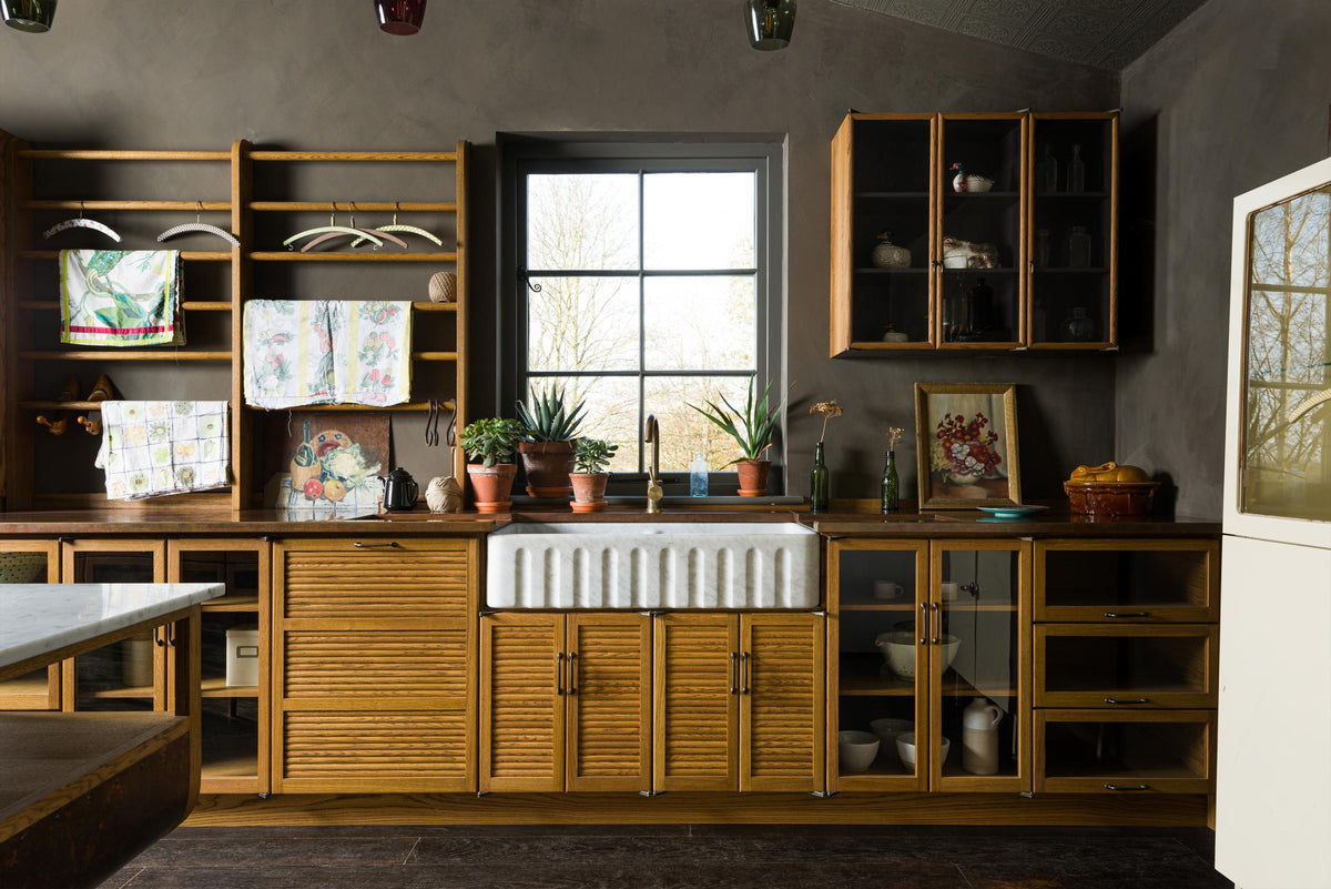 A closer look at a warm and inviting Haberdasher’s kitchen features a long sink run with a copper worktop, a double-fluted marble sink, a kitchen island, and wall cupboards. Adorned with luscious green potted plants, vintage items, artisan ceramics, illustrative tea towels, and paintings, this kitchen creates a lovely exhibitionist space that is both beautiful and practical.